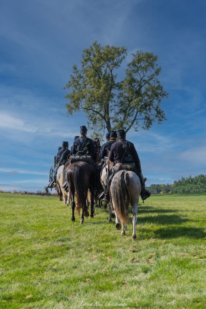 Cavalry soldiers in two rows, riding steadily towards a tree in the distance. Their horses are in motion, and the soldiers are dressed in period uniforms, exuding a sense of purpose and unity as they advance across the open terrain.